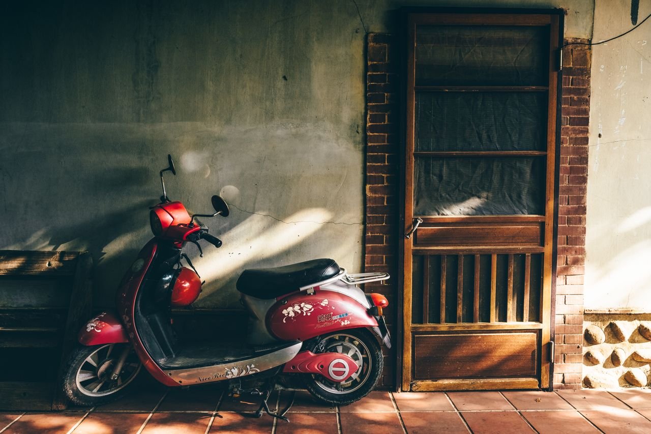 about-01 A vintage red scooter parked next to a wooden door in warm sunlight, casting shadows on a rustic wall.