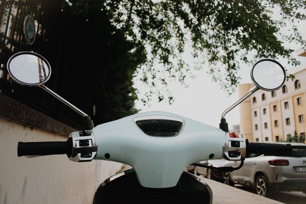 gallery-1 Close-up view of a scooter handlebar and mirrors against an urban backdrop with buildings and greenery.