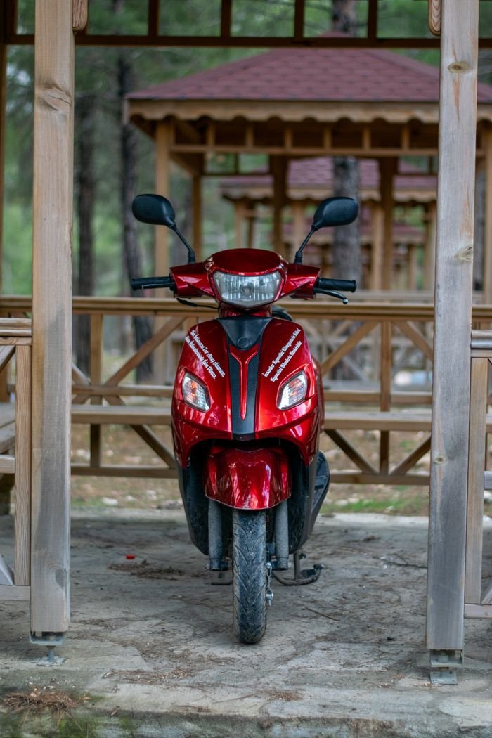 gallery-3 A vibrant red scooter parked in a rustic wooden gazebo in a forest setting.