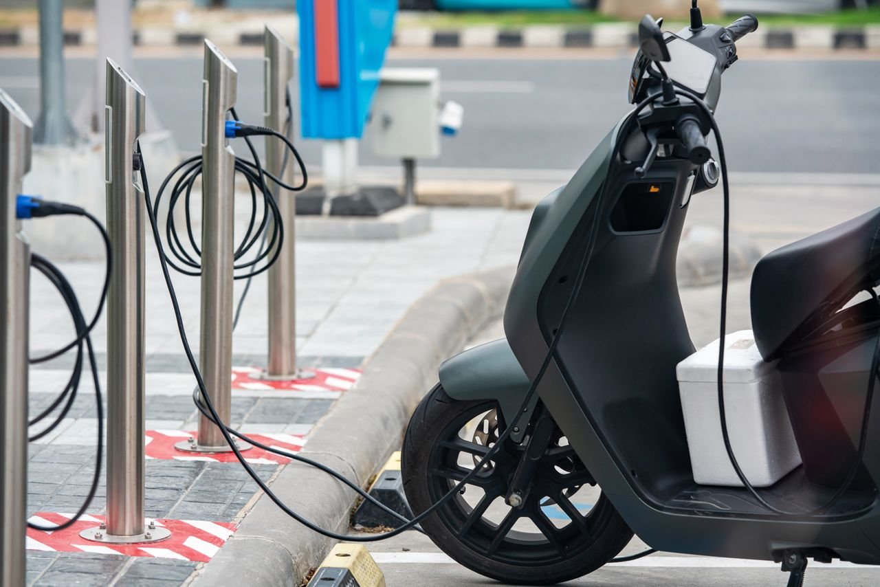 hero-img-01 Electric scooter plugged into a charging station in a city environment, showcasing eco-friendly urban transportation.